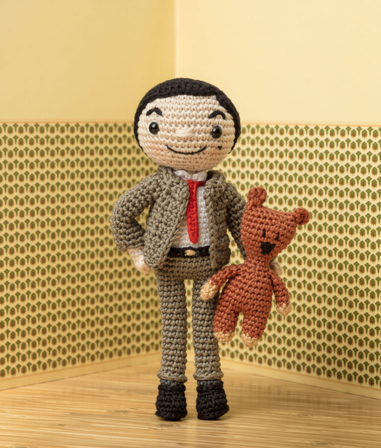 inszeniertes Foto einer Mr.Beann Häkelpuppe vor einer tapezierten Wand mit Teddy im Arm für ein Promi-Häkelbuch