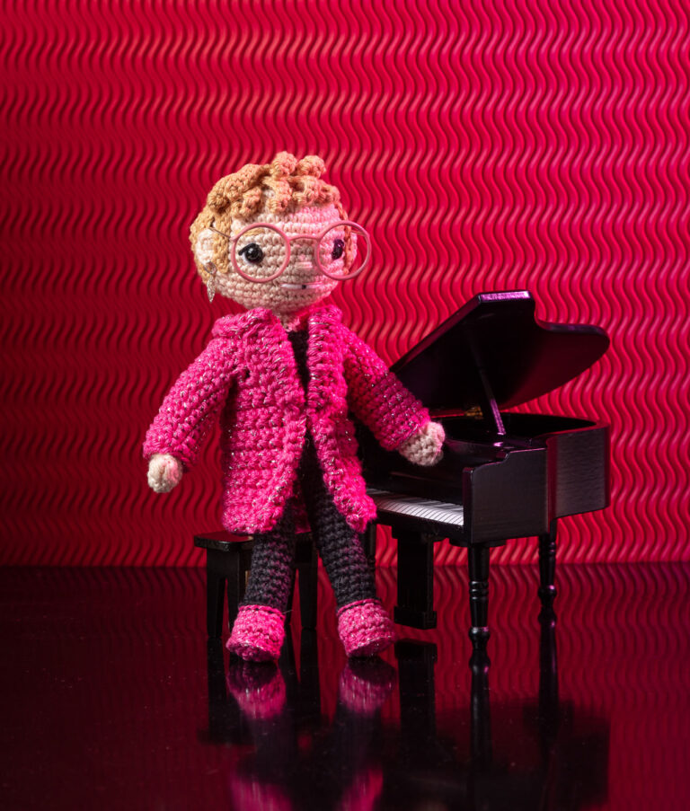 inszeniertes Foto einer Elton John Häkelpuppe vor einem Flügel auf einer Bühne mit pinkem Hintergrund für ein Promi-Häkelbuch