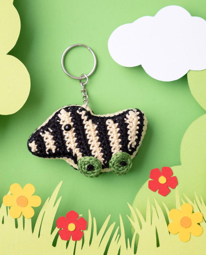 Produktfoto einer DIY gehäkelten Tigerente als Schlüsselanhänger in einer Paper-Art-Kulisse mit Grün, Wolken und Blumen