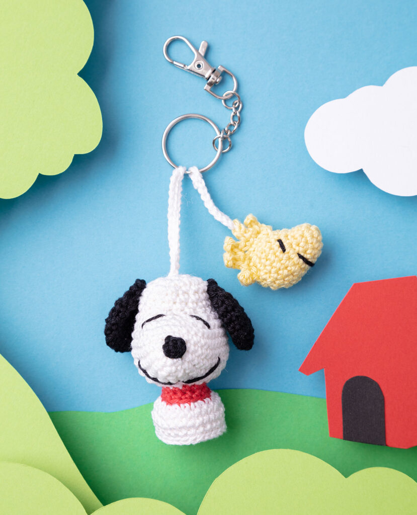 Produktfoto eines DIY gehäkelten Snoopy als Schlüsselanhänger in einer Paper-Art-Kulisse mit Grün, Wolken und Hundehütte