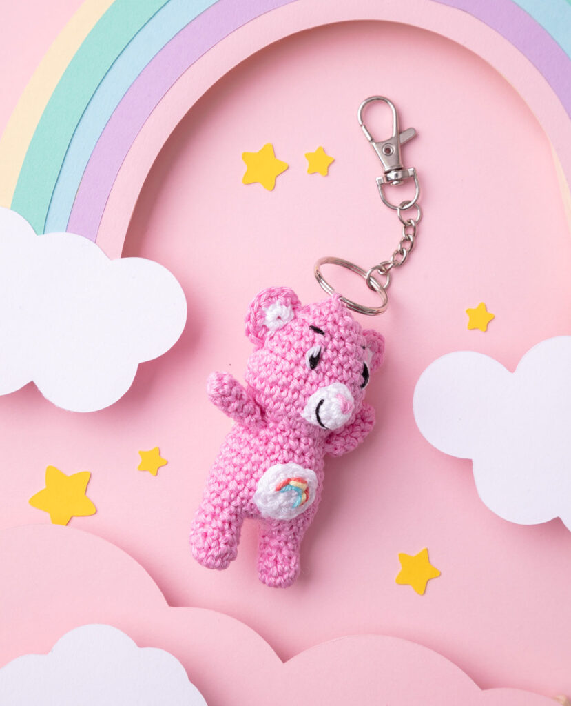 Produktfoto eines DIY gehäkelten Glücksbärchens als Schlüsselanhänger in einer Paper-Art-Kulisse auf Rosa mit einem Regenbogen, Wolken und Sternen