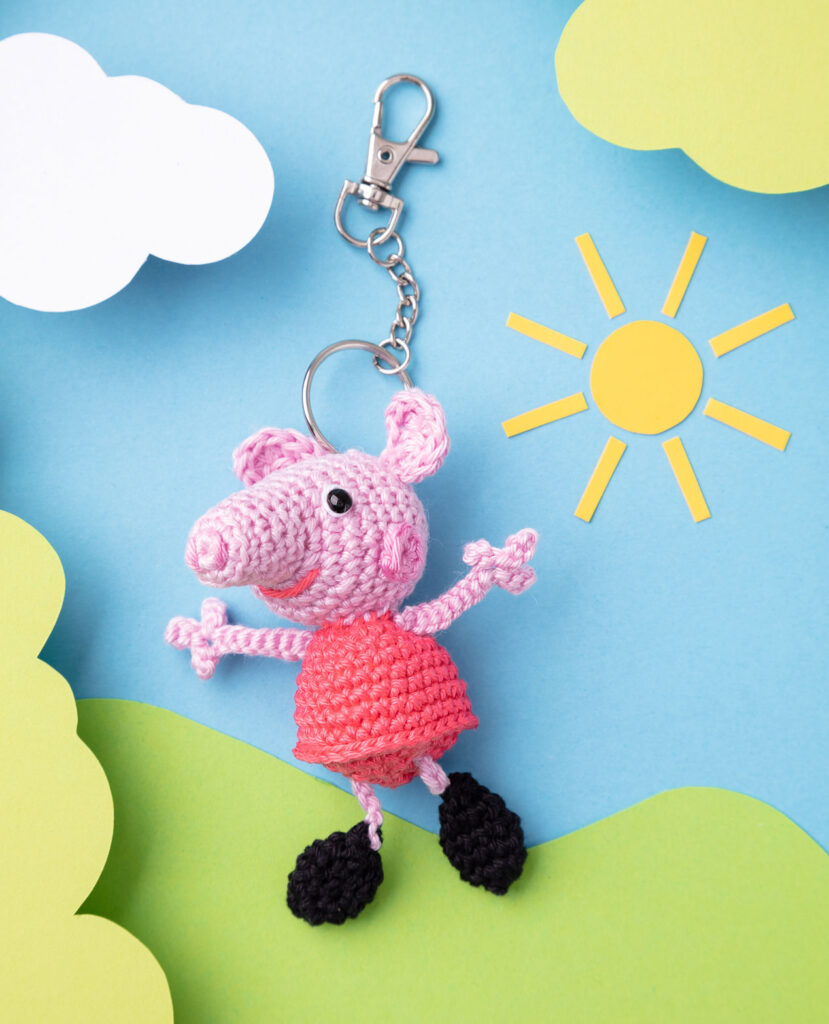 Produktfoto einer DIY gehäkelten Peppa Wutz als Schlüsselanhänger in einer Paper-Art-Kulisse mit Grün, Wolken und Sonne