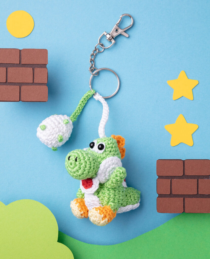 Produktfoto eines DIY gehäkelten Yoshi als Schlüsselanhänger in einer dem Computerspiel nachempfundenen Paper-Art-Kulisse