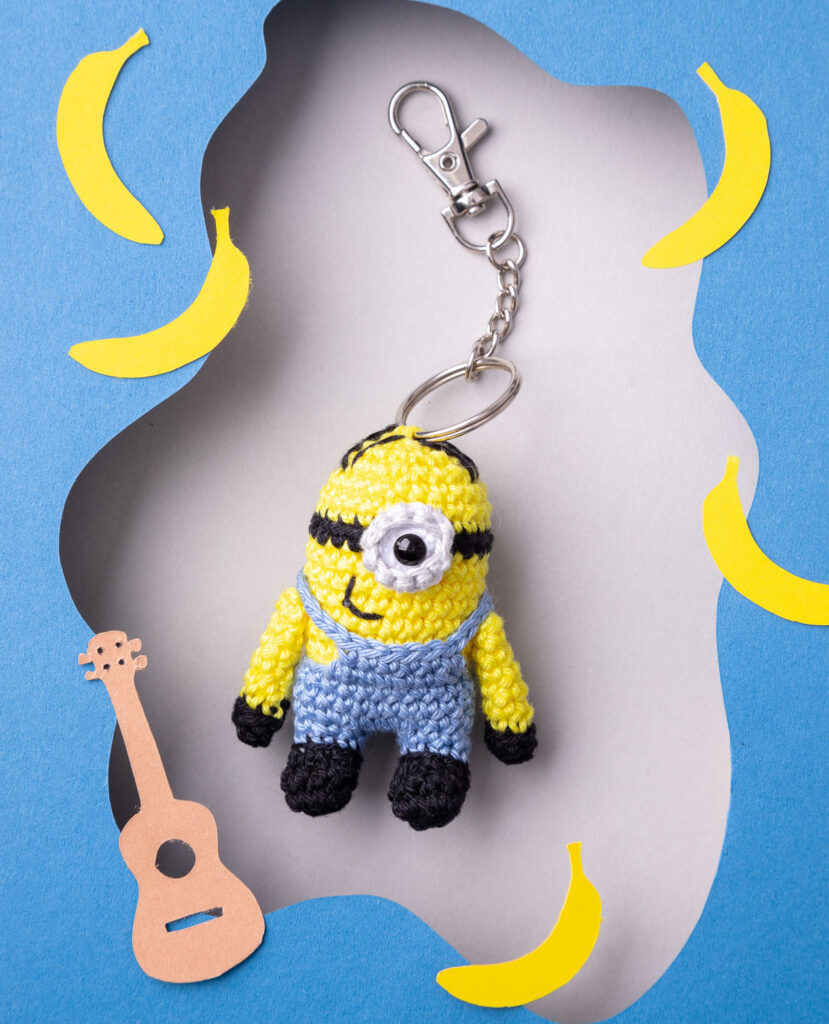 Produktfoto eines DIY gehäkelten Minion als Schlüsselanhänger in einer Paper-Art-Kulisse mit blau, Gitarre und Bananen