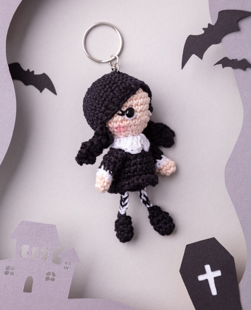 Produktfoto einer DIY gehäkelten Wednesday Adams als Schlüsselanhänger in einer Paper-Art-Kulisse mit dunkelgrau, Fledermäusen, ihrem Zuhause und einem kleinen Sarg