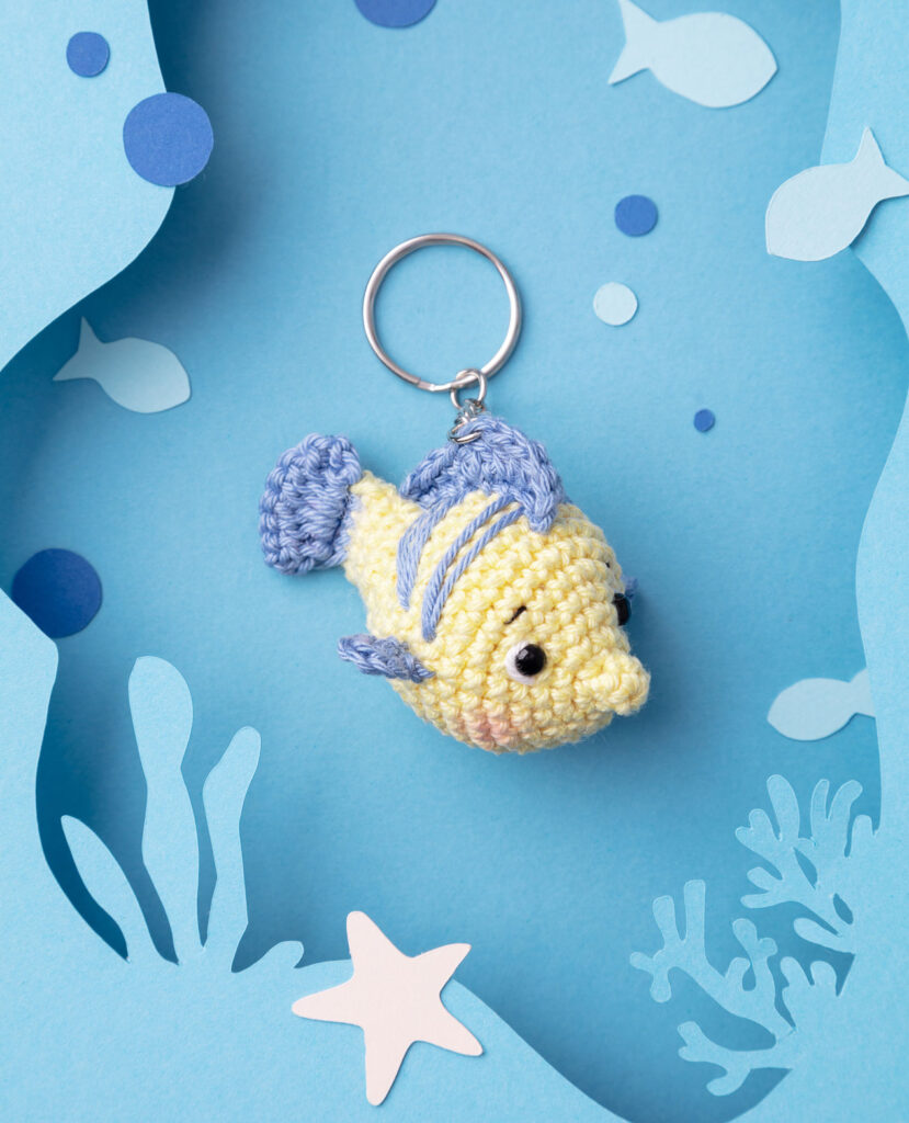 Produktfoto eines DIY gehäkelten Fabius aus Arielle als Schlüsselanhänger in einer Paper-Art-Kulisse mit blau, Fischen, Meeresalgen und Luftblasen