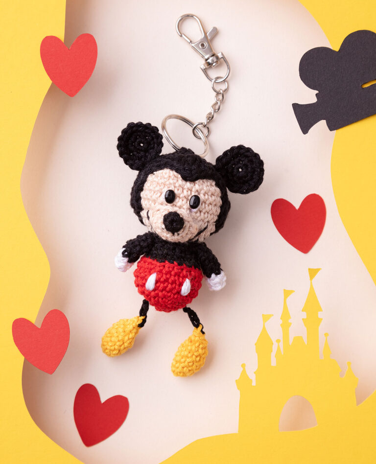 Produktfoto einer DIY gehäkelten Mickey Maus als Schlüsselanhänger in einer Paper-Art-Kulisse mit Gelb, dem Disneyschloss, roten Herzen und einer Kamera