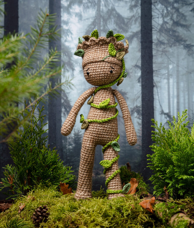 Produktfoto einer Häkelpuppe verkleidet als Superheld in einem aufwendigen Setting das einen düsteren Wald zeigt, dekoriert mit echten Naturmaterialien wie Zweige, Moos und Zapfen