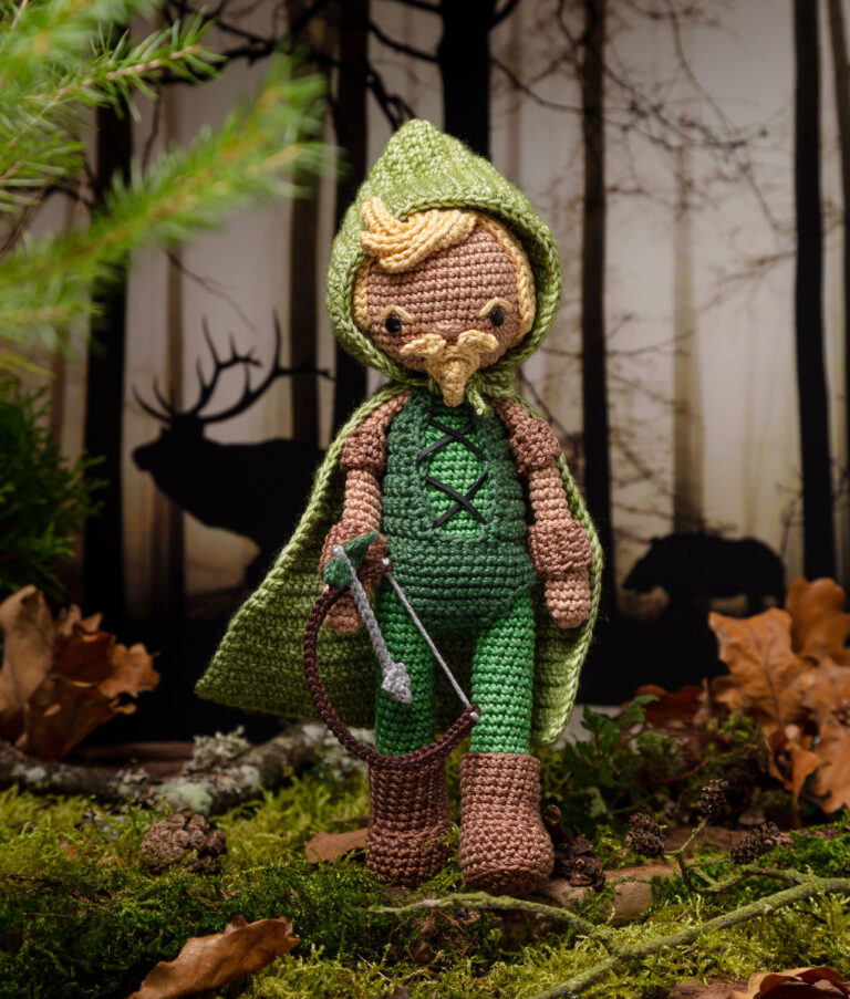Produktfoto einer Häkelpuppe verkleidet als Superheld mit Pfeil und Bogen in einem aufwendigen Setting das einen düsteren Wald zeigt, dekoriert mit echten Naturmaterialien wie Zweige, Moos und Zapfen