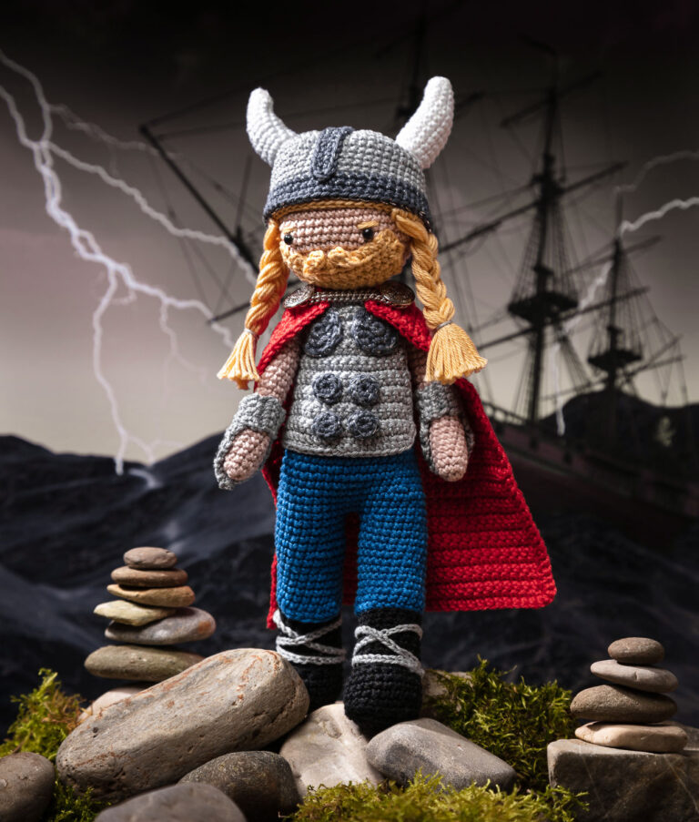 Produktfoto einer Häkelpuppe verkleidet als Wikinger-Superheld in einem aufwendigen Setting das eine düstere Szene auf See zeigt zeigt, dekoriert mit echten Naturmaterialien wie Steine und Moos