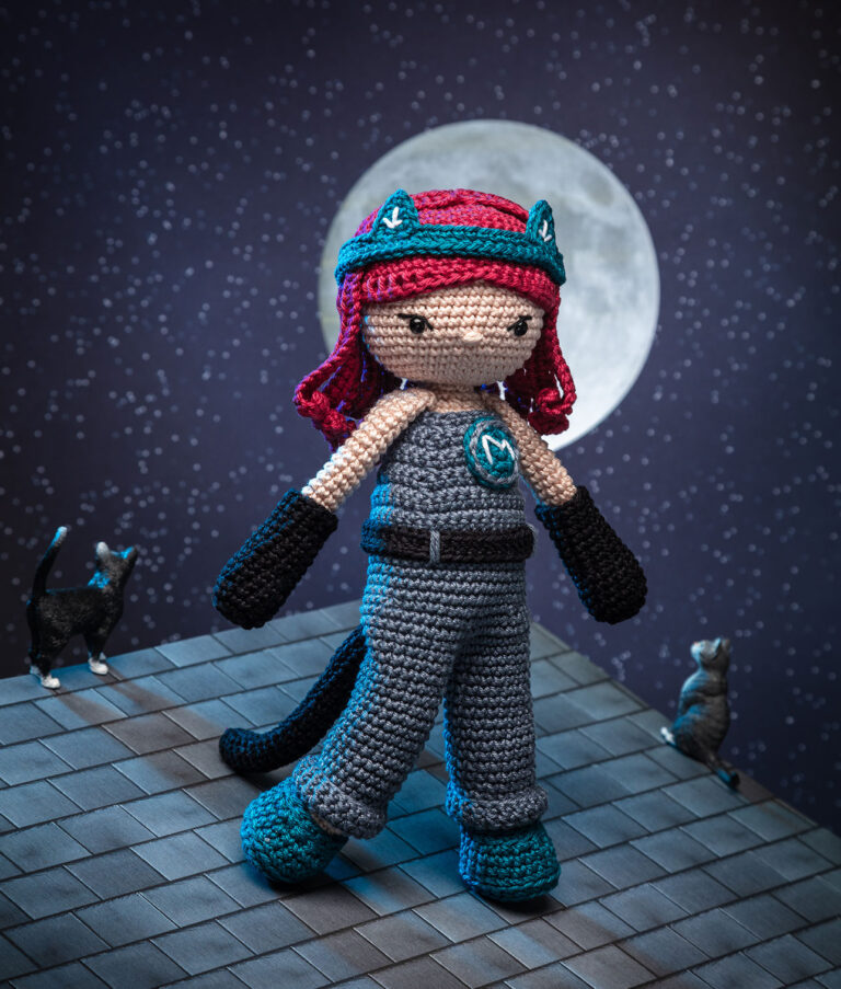 Produktfoto einer Häkelpuppe verkleidet als geheimnisvolle Superheldin in einem aufwendigen Setting das ein Häuserdach vor einem Nachhimmel zeigt, mit Vollmond und zwei Katzen