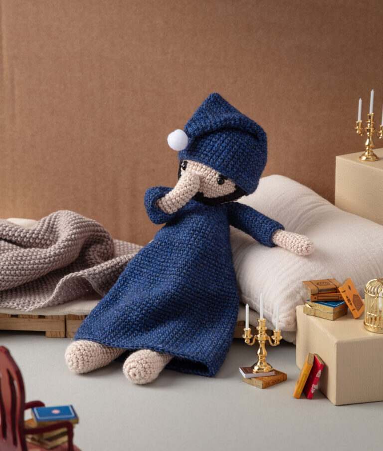 Produktfoto einer Häkelpuppe verkleidet als arme Poet in einem aufwendigen Setting das einen Dachboden mit Bett, Büchern und Kerzenleuchtern zeigt