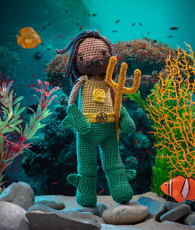 Produktfoto einer Häkelpuppe verkleidet als Unterwasser-Superheld mit Dreizack in einem aufwendigen Setting das eine düstere Unterwasserwelt zeigt, dekoriert mit Sand, Steinen und Wasserpflanzen sowie zwei Fischen