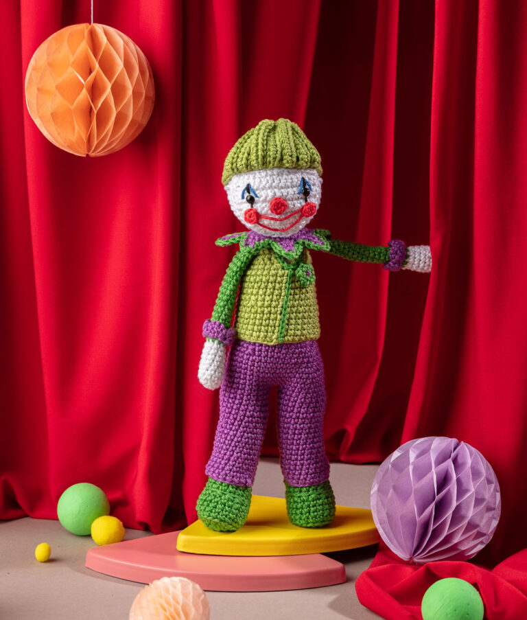 Produktfoto einer Clown-Häkelpuppe in einem aufwendigen Zirkus-Setting mit rotem Vorhang, Wabenbällen und bunten Bällen am Boden