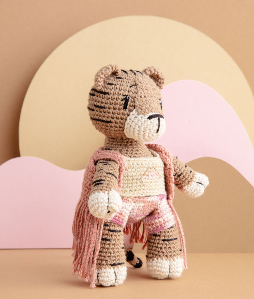 inszeniertes Produktfoto einer bekleideten Tiger Häkelpuppe vor einem Paper-Art Hintergrund mit verschiedenfarbigen runden, organischen Formen für ein Häkelbuch
