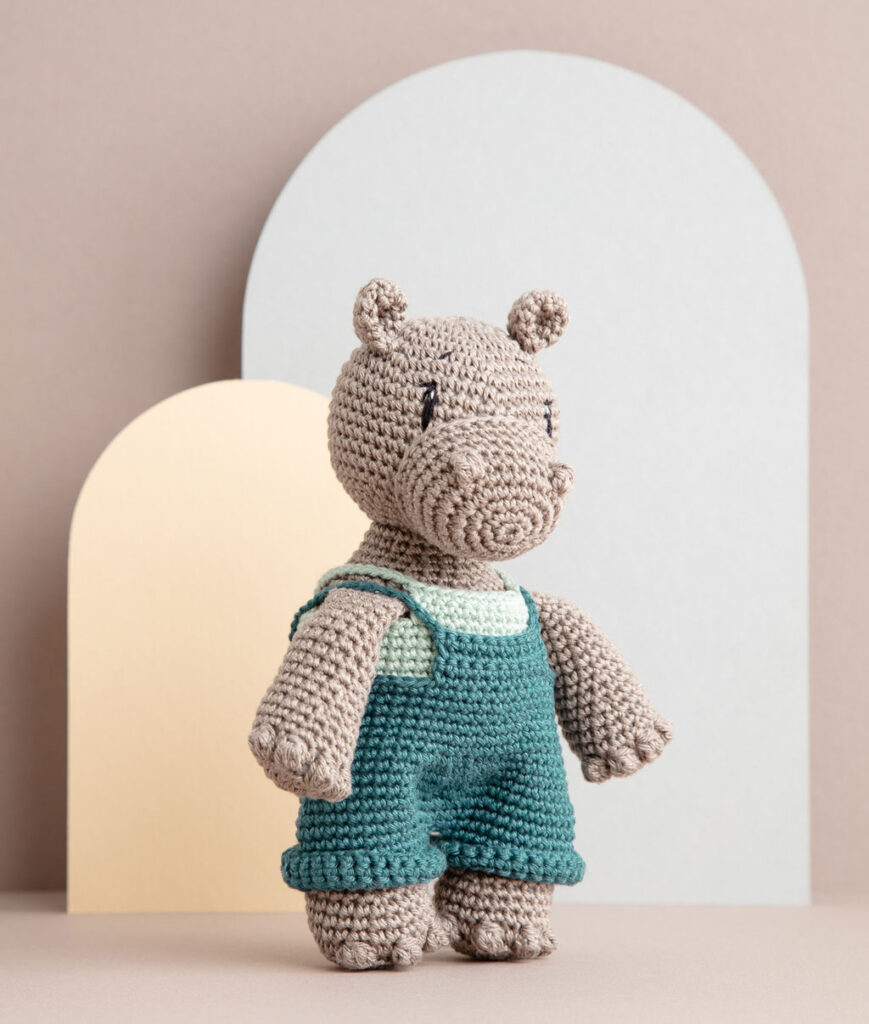 inszeniertes Produktfoto einer bekleideten Nilpferd-Häkelpuppe vor einem Paper-Art Hintergrund mit verschiedenfarbigen runden, organischen Formen für ein Häkelbuch