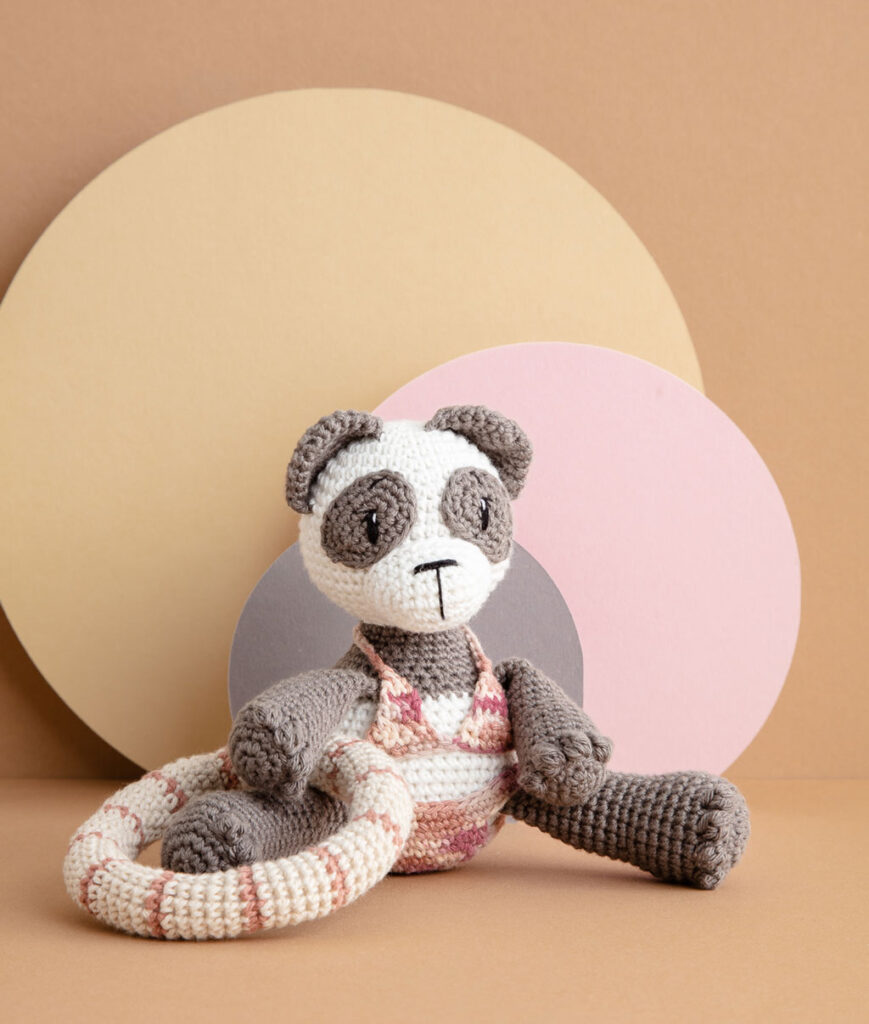 inszeniertes Produktfoto einer bekleideten Pandabär-Häkelpuppe vor einem Paper-Art Hintergrund mit verschiedenfarbigen runden, organischen Formen für ein Häkelbuch
