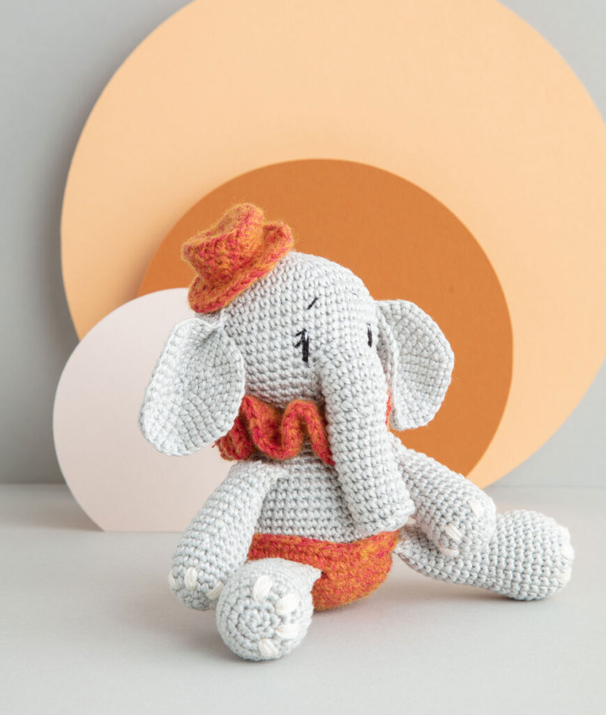 inszeniertes Produktfoto einer bekleideten Elefanten-Häkelpuppe vor einem Paper-Art Hintergrund mit verschiedenfarbigen runden, organischen Formen für ein Häkelbuch