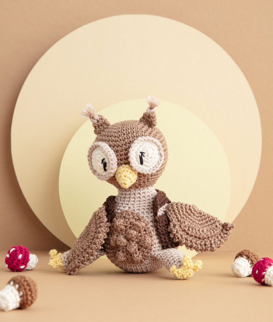 inszeniertes Produktfoto einer bekleideten Eulen-Häkelpuppe vor einem Paper-Art Hintergrund mit verschiedenfarbigen runden, organischen Formen für ein Häkelbuch
