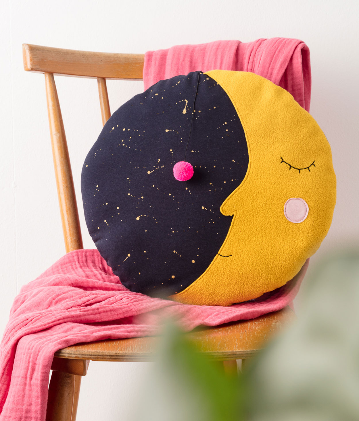 Produktfoto eines selbstgenähten Mond-Kissens auf einem Holzstuhl, dekoriert mit einer pinkfarbenen Decke