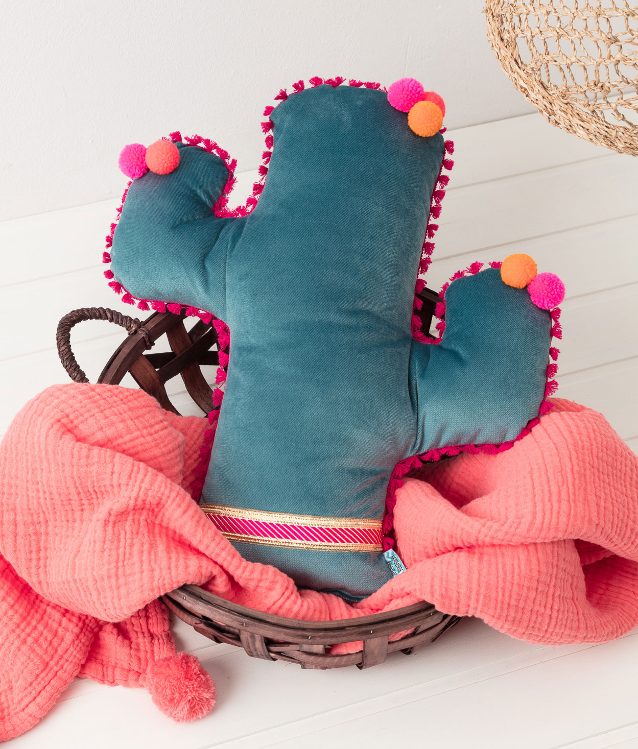 Produktfoto eines selbstgenähten Kaktus-Kissens in einem Körbchen in einer Interior-Kulisse mit einer pinkfarbenen Decke und Lampenschirm