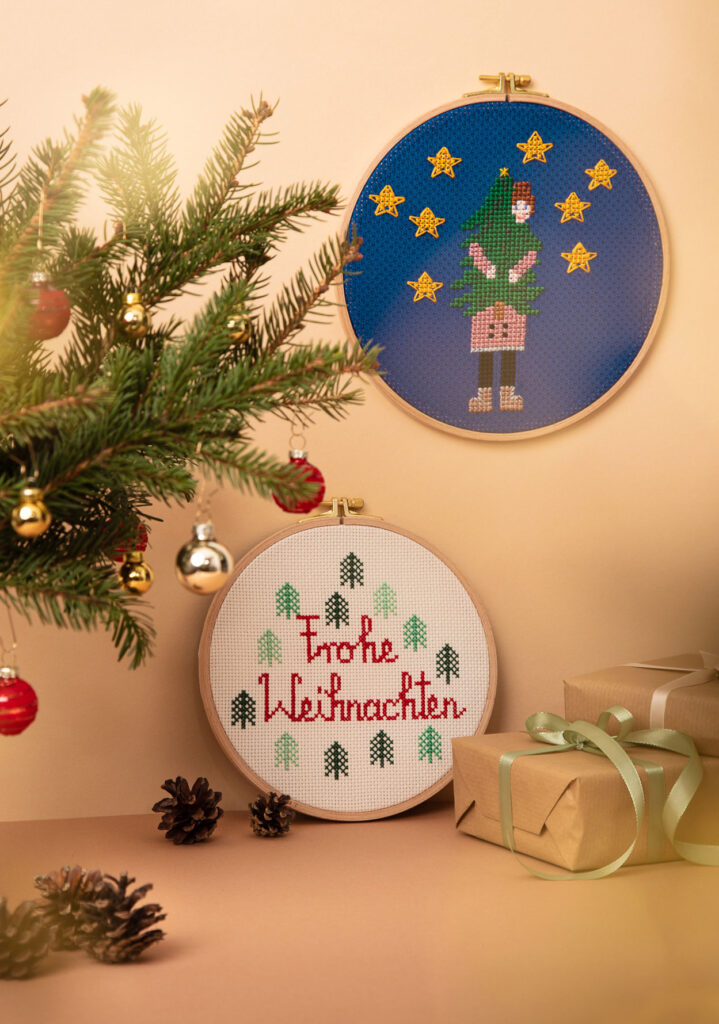 weihnachtlich inszeniertes Produktfoto von zwei bestickten Stickrahmen an einer Wand hängend, die zwei weihnachtliche Motive zeigen, zwei Geschenke, Tannenzweige mit Kugeln und Tannenzapfen dienen als Kulisse
