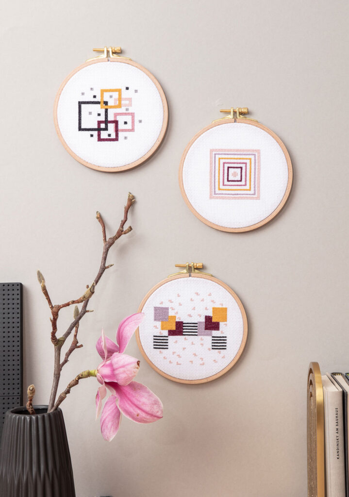 Produktfoto von drei bestickten Stickrahmen an einer grauen Wand hängend, die grafische Muster zeigen, Bücher und eine Vase mit Magnolien dienen als Kulisse