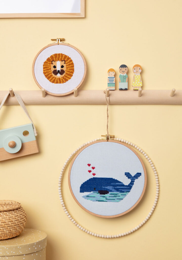 Produktfoto von zwei bestickten Stickrahmen - ein Löwe und ein Wal - die an einer Hakenleiste eines Kinderzimmers drapiert sind, Bilderrahmen, Holzfiguren und Deko-Boxen dienen als Foto-Kulisse