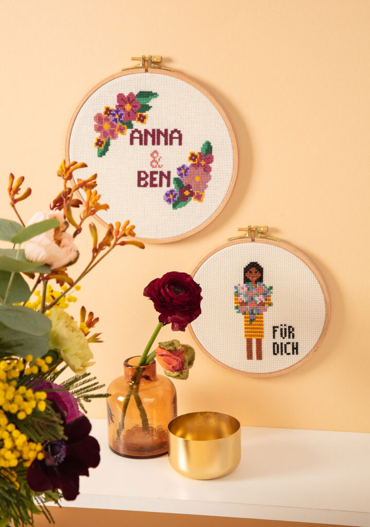 gemütlich inszeniertes Produktfoto von zwei bestickten Stickrahmen an einer Wand hängend, die zwei Motive mit Blumen zeigen, Vasen mit Blumen dienen als Dekoration der Foto-Kulisse