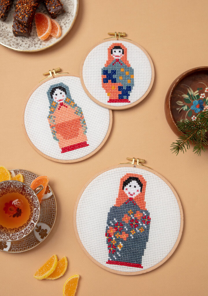 gemütlich inszeniertes Produktfoto von drei bestickten Stickrahmen die drei Matroschkas zeigen, Teetassen, russische Süßigkeiten und eine Folklore-Schale dienen als Dekoration