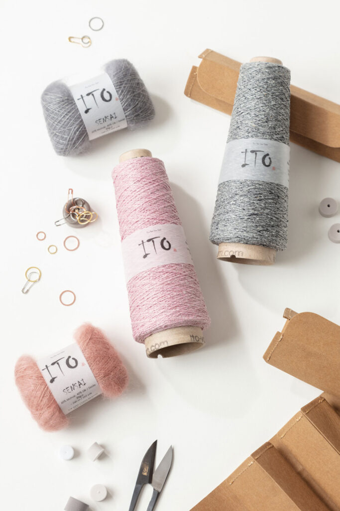 Produktfoto zum Thema Stricken mit mehreren Knäuel und Konen der Firma ITO, Stricknadeln und weiteren Strickutensilien in den Farben grau, rosa und braun