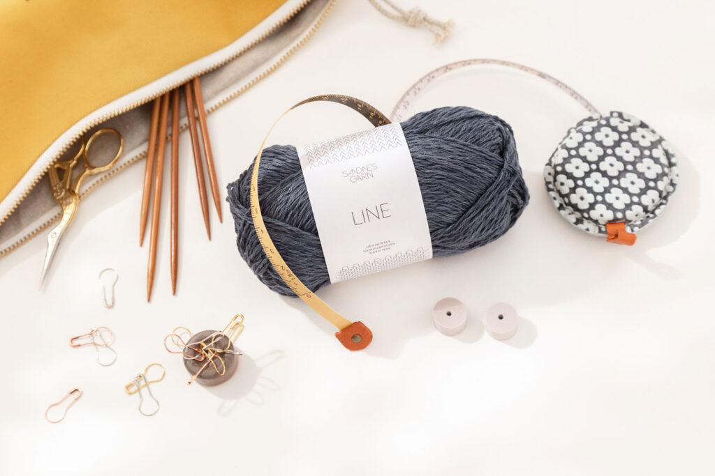Produktfoto eines schönen Arrangements zum Thema Stricken mit einem blauen Wollknäuel von Sandnes Garn, verschiedenen Stricknadeln in einer gelben Projekttasche und verschiedenem Strickzubehör
