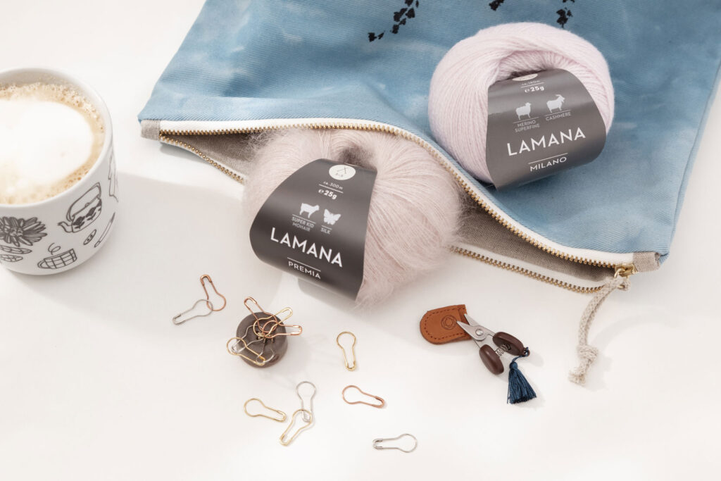 Produktfoto eines schönen Arrangements zum Thema Stricken mit zwei rosanen Wollknäuel von Lamana, einer blauen Projekttasche, einer Kaffeetasse und verschiedenem Strickzubehör