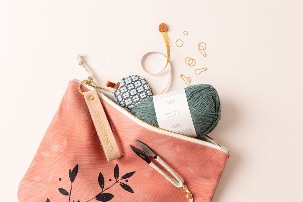 Produktfoto eines dynamisch fotografierten Arrangements zum Thema Stricken mit einer roten Projekttasche aus der ein Wollknäuel von Sandnes Garn herausschaut, ein Maßband und verschiedenes Strickzubehör