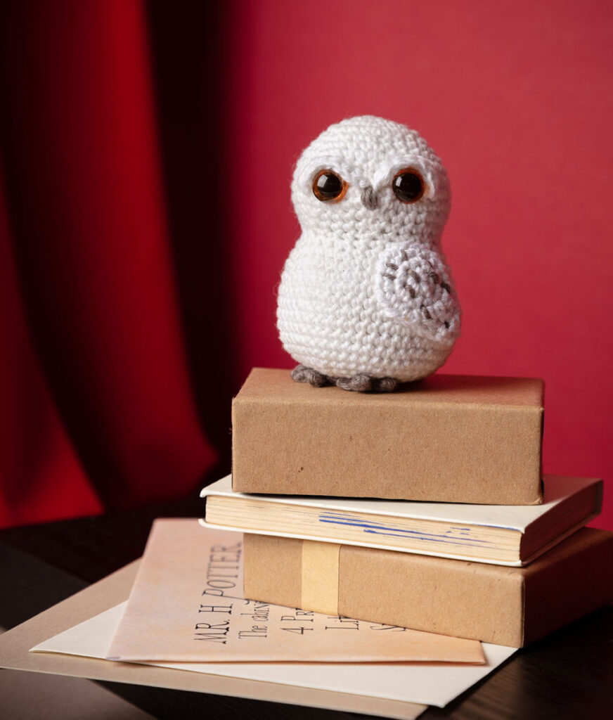 Produktfoto einer Hedvig Eulen-Häkelfigur aus Harry Potter in Mini-Kulisse mit rotem Vorhang, Paketen und Briefen