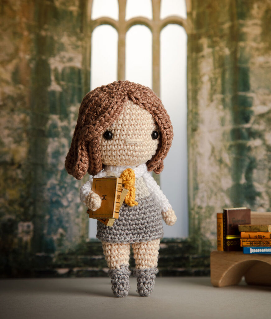 Produktfoto einer Hermine Häkelfigur aus Harry Potter in nachgestellter Mini-Kulisse eines Raums von Hogwarts mit Büchern im Arm