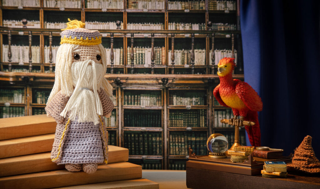 Produktfoto einer Dumbledore Häkelfigur aus Harry Potter in nachgestellter Mini-Kulisse seiner Bibliothek mit Fawkes, Zauberkugel und dem sprechenden Hut