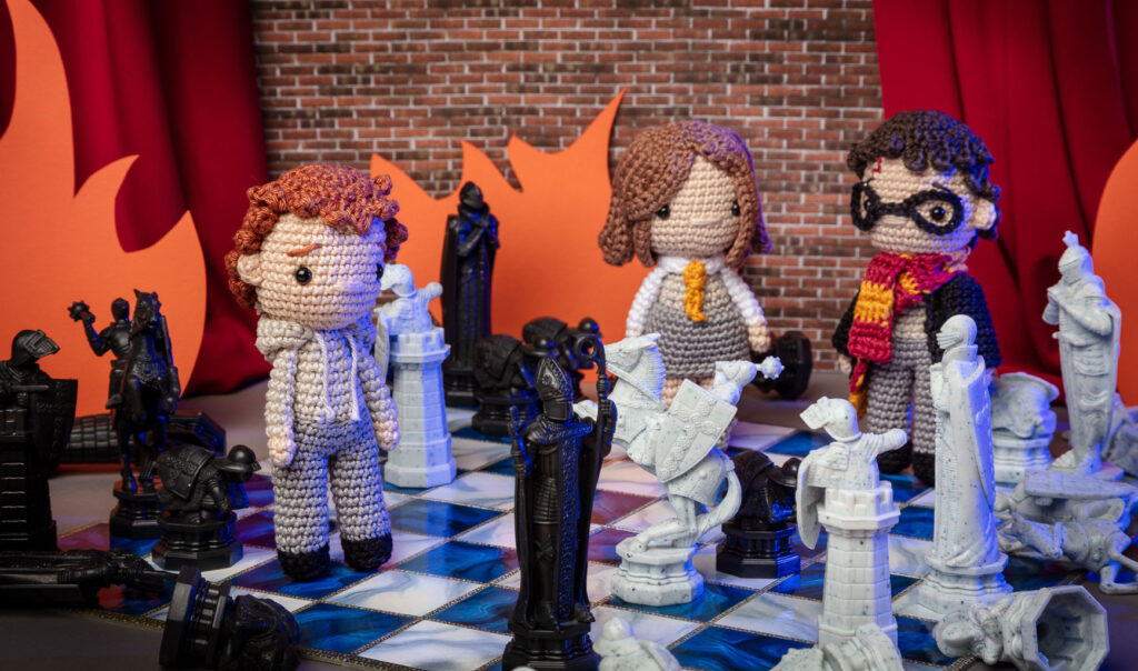Produktfoto von drei Häkelfiguren - Harry Potter, Hermine & Ron - in nachgestellter Mini-Kulisse der Zauberschach-Szene aus Harry Potter
