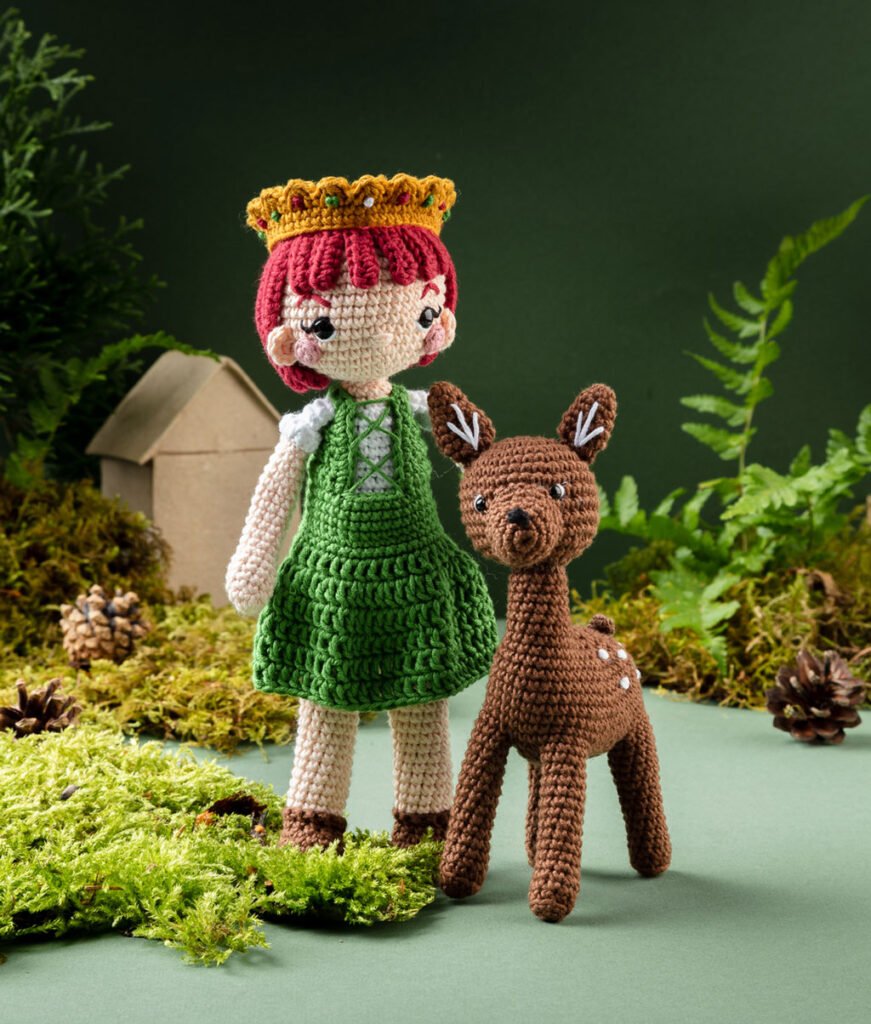 Produktfoto von zwei modern gestalteten Häkelfiguren - eines Mädchens mit Krone und eines Rehs aus dem Märchen Brüderchen und Schwesterchen - in einer dunklen Kulisse aus Papier dekoriert mit Zweigen, Moos, Zapfen und einem kleinen Häuschen