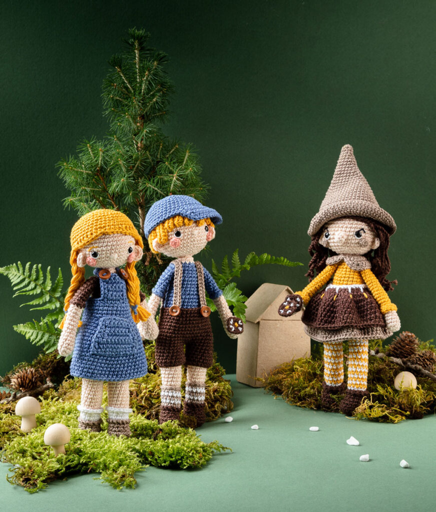 Produktfoto von drei modern gestalteten Häkelfiguren - Hänsel, Gretel und einer Hexe - in einer dunklen Kulisse aus Papier dekoriert mit Zweigen, Farne und Moos
