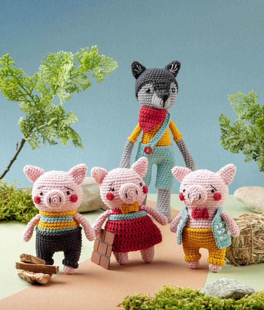 Produktfoto von vier modern gestalteten Häkelfiguren - drei Schweinchen und einem Wolf aus dem Märchen die drei Schweinchen - in einer Kulisse aus Papier dekoriert mit Zweigen, Steine und Moos