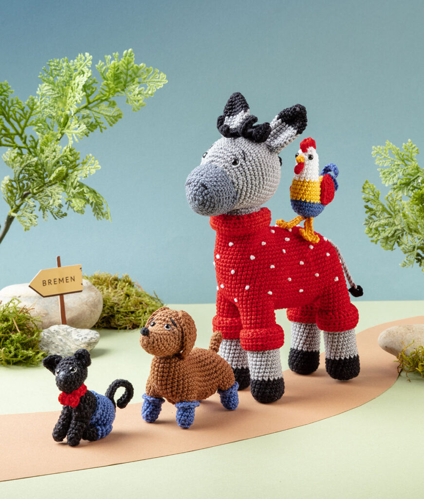 Produktfoto von vier modern gestalteten Häkelfiguren - Esel, Hahn, Hund und Katze aus den Bremer Stadtmusikanten - in einer Kulisse aus Papier dekoriert mit Zweigen, Steine und Moos