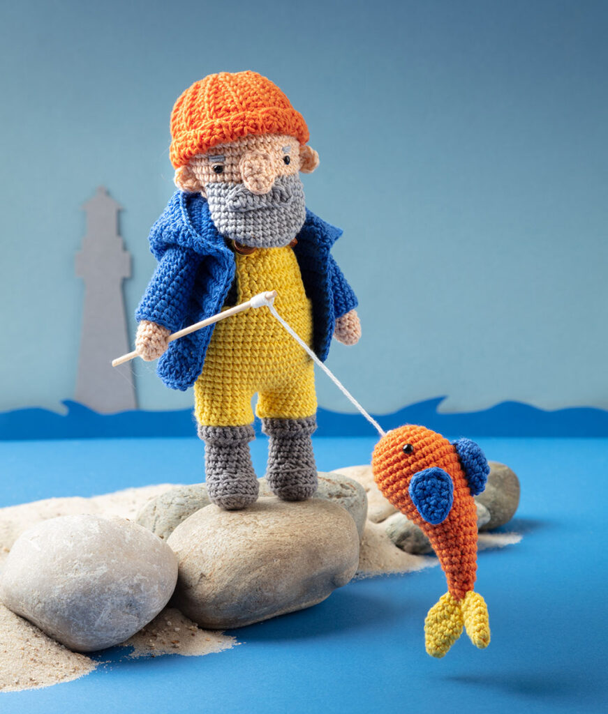 Produktfoto der gehäkelten Figur eines Fischers aus dem Märchen "Vom Fischer und seiner Frau" in einer Kulisse aus Papier dekoriert mit Naturmaterialien wie Steinen und Sand