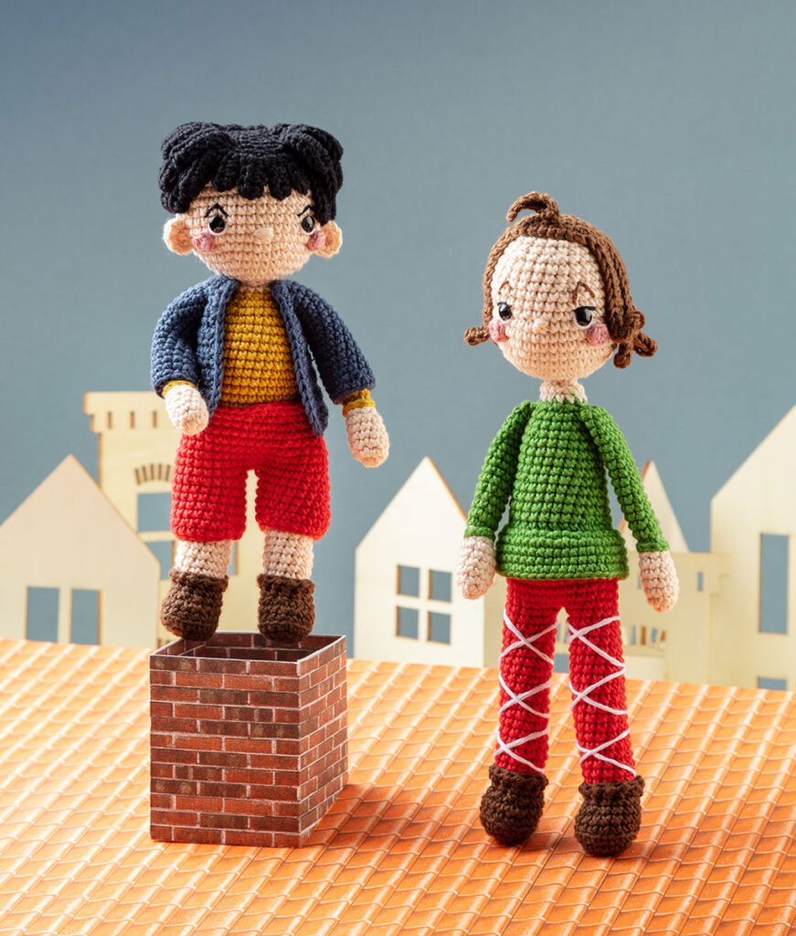 Produktfoto von zwei modern gestalteten Häkelfiguren - Max und Moritz - in einer Kulissen mit Dach, Schornstein und Häusern im Hintergrund