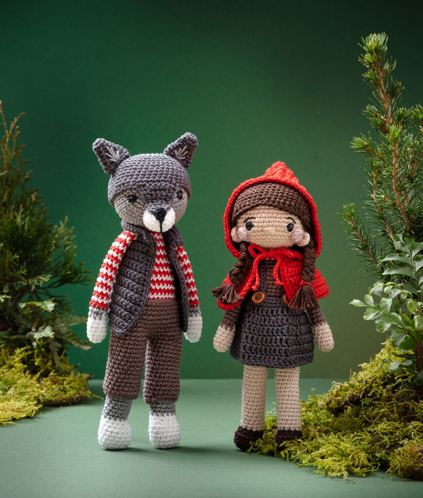 Produktfoto von zwei modern gestalteten Häkelfiguren - Rotkäppchen und der Wolf - in einer grünen Kulisse dekoriert mit Naturmaterialien wie Tannenbäumchen, Farne und Moos
