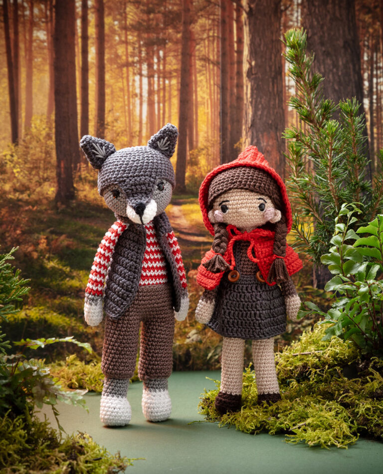 Titelfoto von zwei modern gestalteten Häkelfiguren - Rotkäppchen und der Wolf - in einer gestalteten Kulisse dekoriert mit Naturmaterialien wie Tannenbäumchen, Farne und Moos