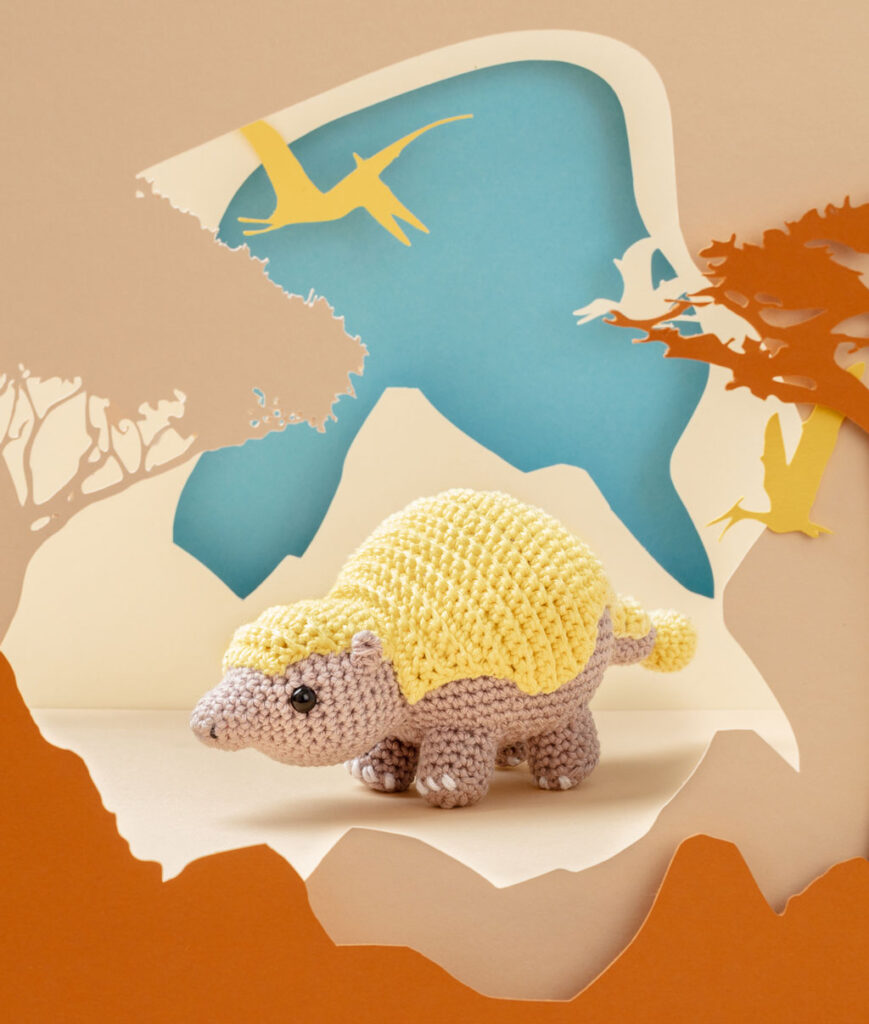 Produktfoto eines gelb-beigen Dinosauriers in beiger Paper-Art Kulisse mit verschiedenen Ebenen, die eine Landschaft der Urzeit ergeben