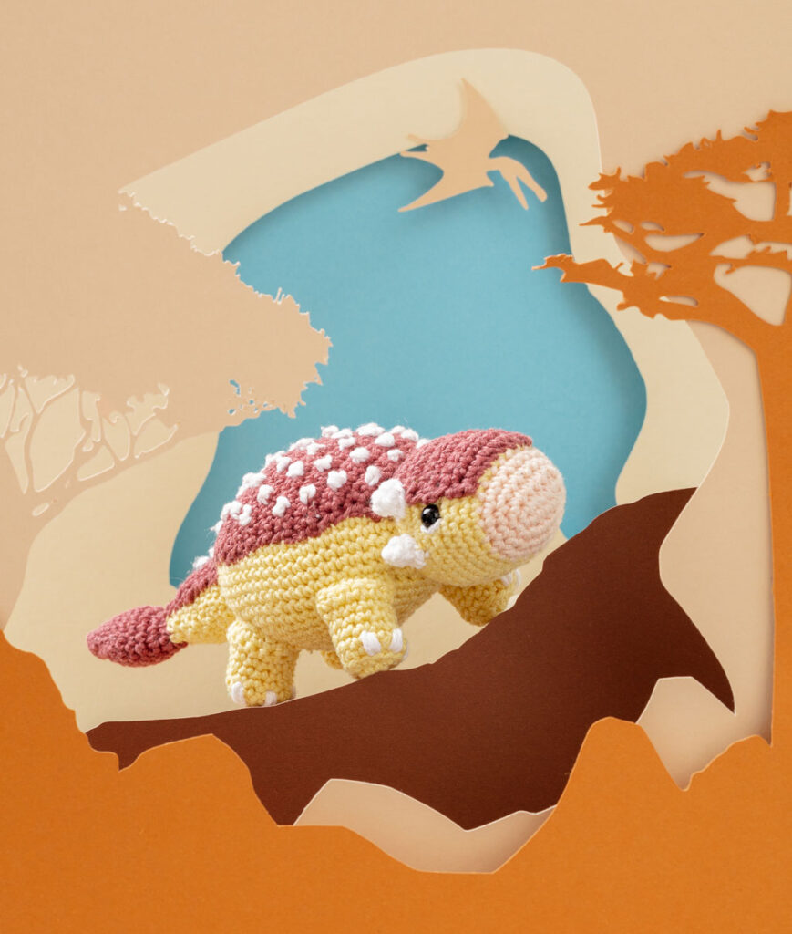 Produktfoto eines gelb-braunen Dinosauriers in beiger Paper-Art Kulisse mit verschiedenen Ebenen, die eine Landschaft der Urzeit ergeben
