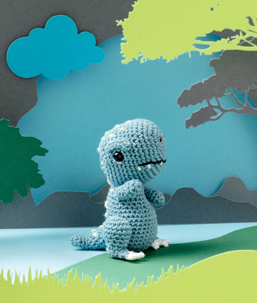 Produktfoto eines blauen Dinosaurier-Babys in blauer Paper-Art Kulisse mit verschiedenen Ebenen, die eine Landschaft der Urzeit ergeben
