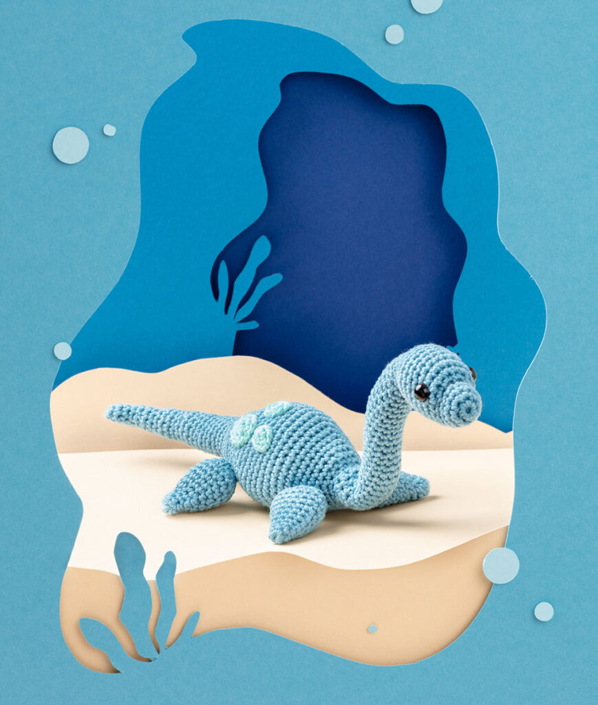 Produktfoto eines blauen Dinosauriers in blauer Paper-Art Kulisse mit verschiedenen Ebenen, die eine Meeres-Landschaft der Urzeit ergeben